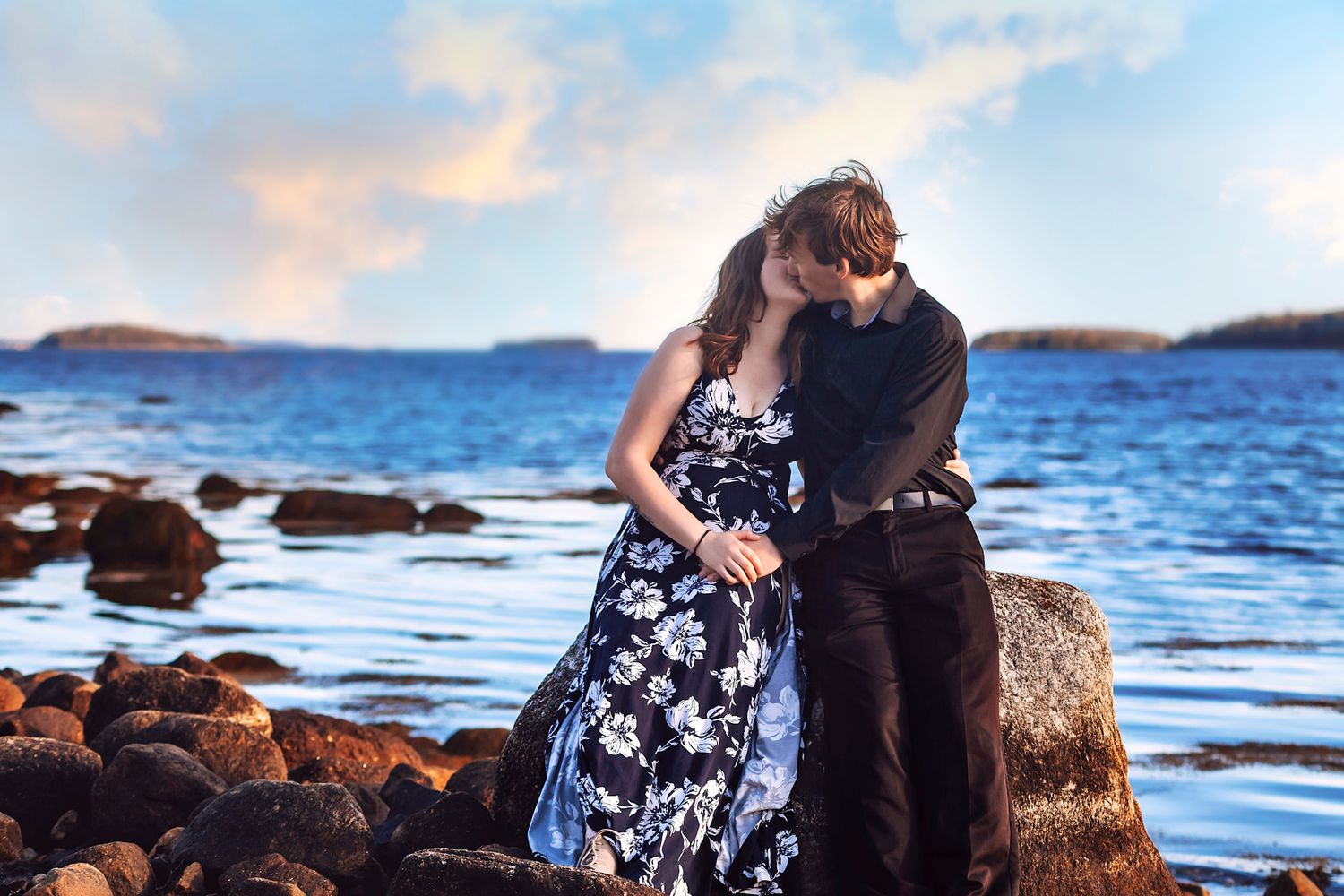 Romantic couple embracing on rocky shoreline at sunset, woman in floral dress, scenic ocean backdrop.