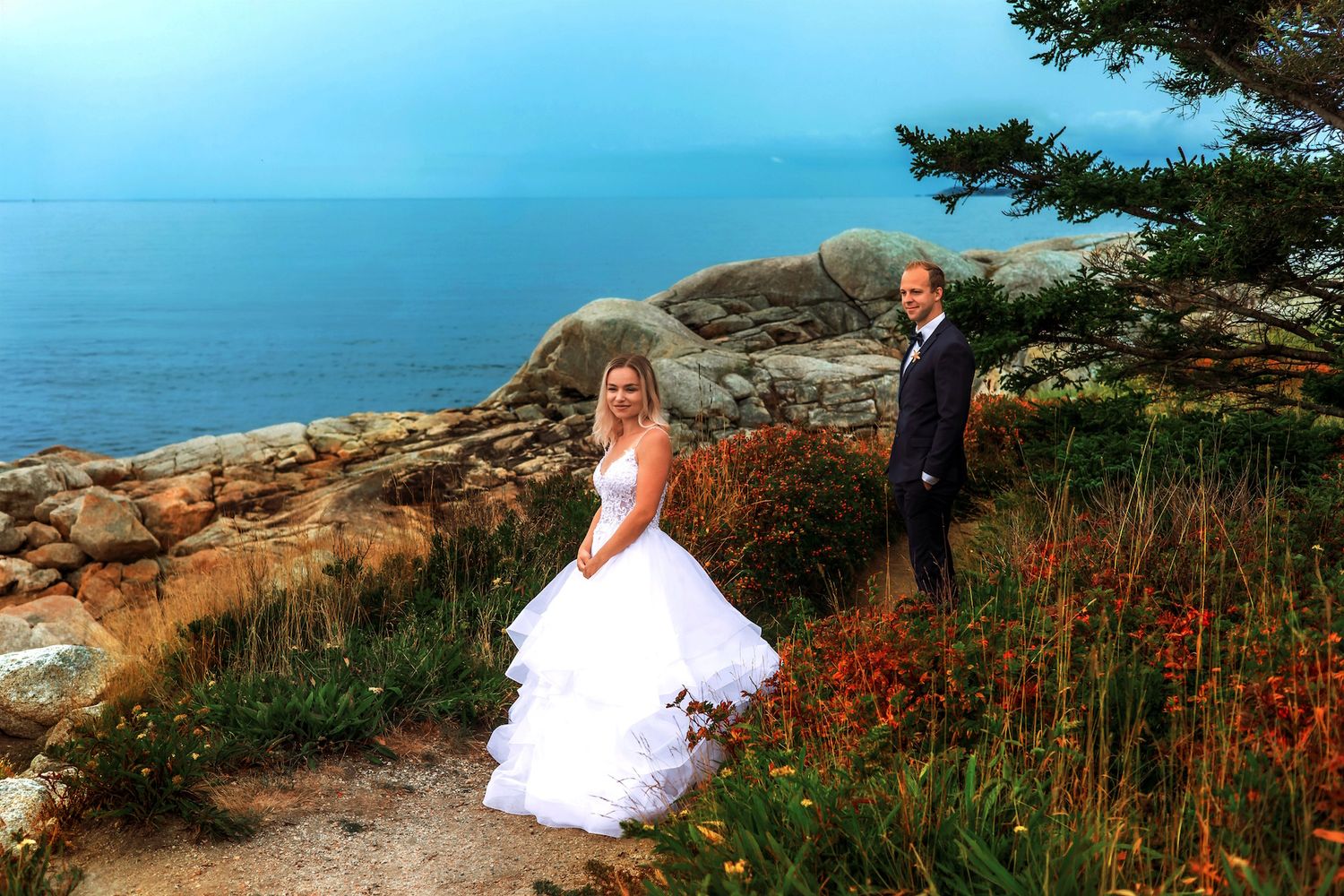 Bride in white gown and groom in suit pose on rocky coastal cliffside with ocean and pine trees in background.