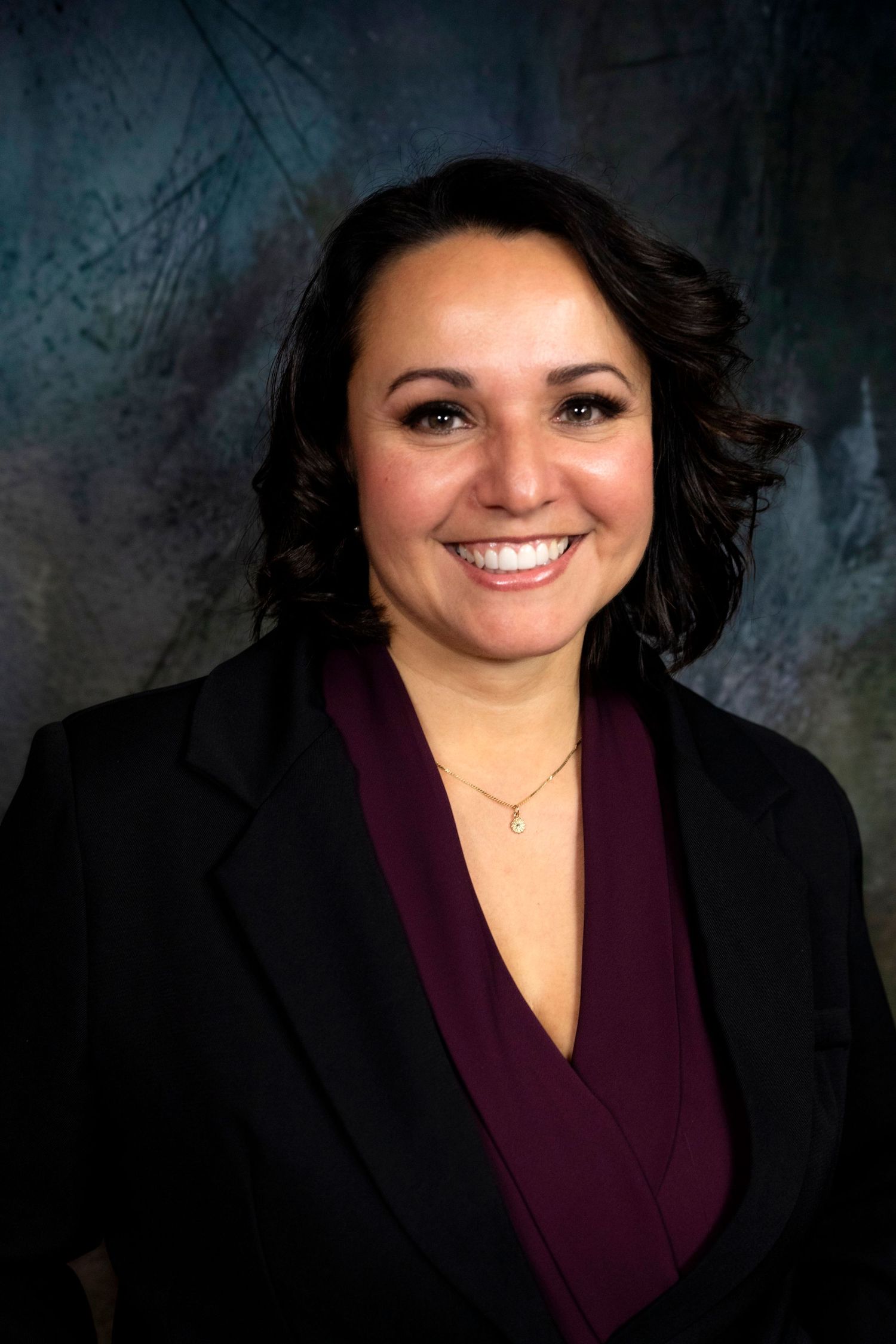 Professional headshot of a smiling woman with dark hair wearing a black blazer and burgundy top.