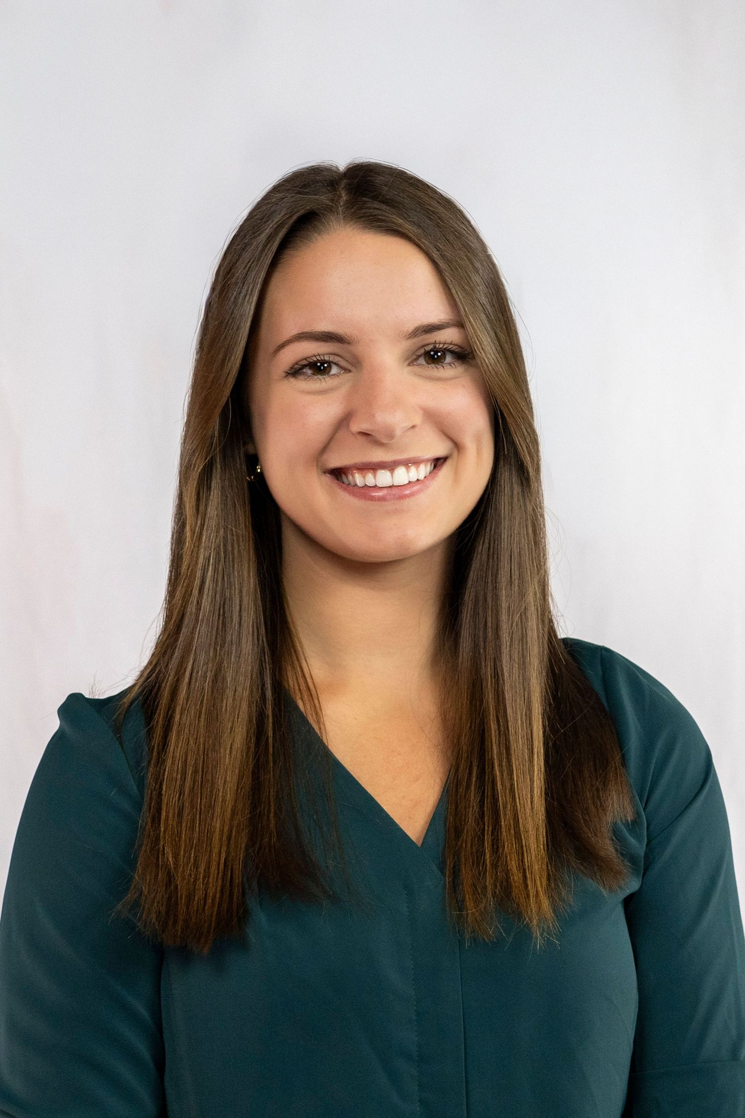 Smiling young woman with long brown hair wearing a teal v-neck top against a white background.
