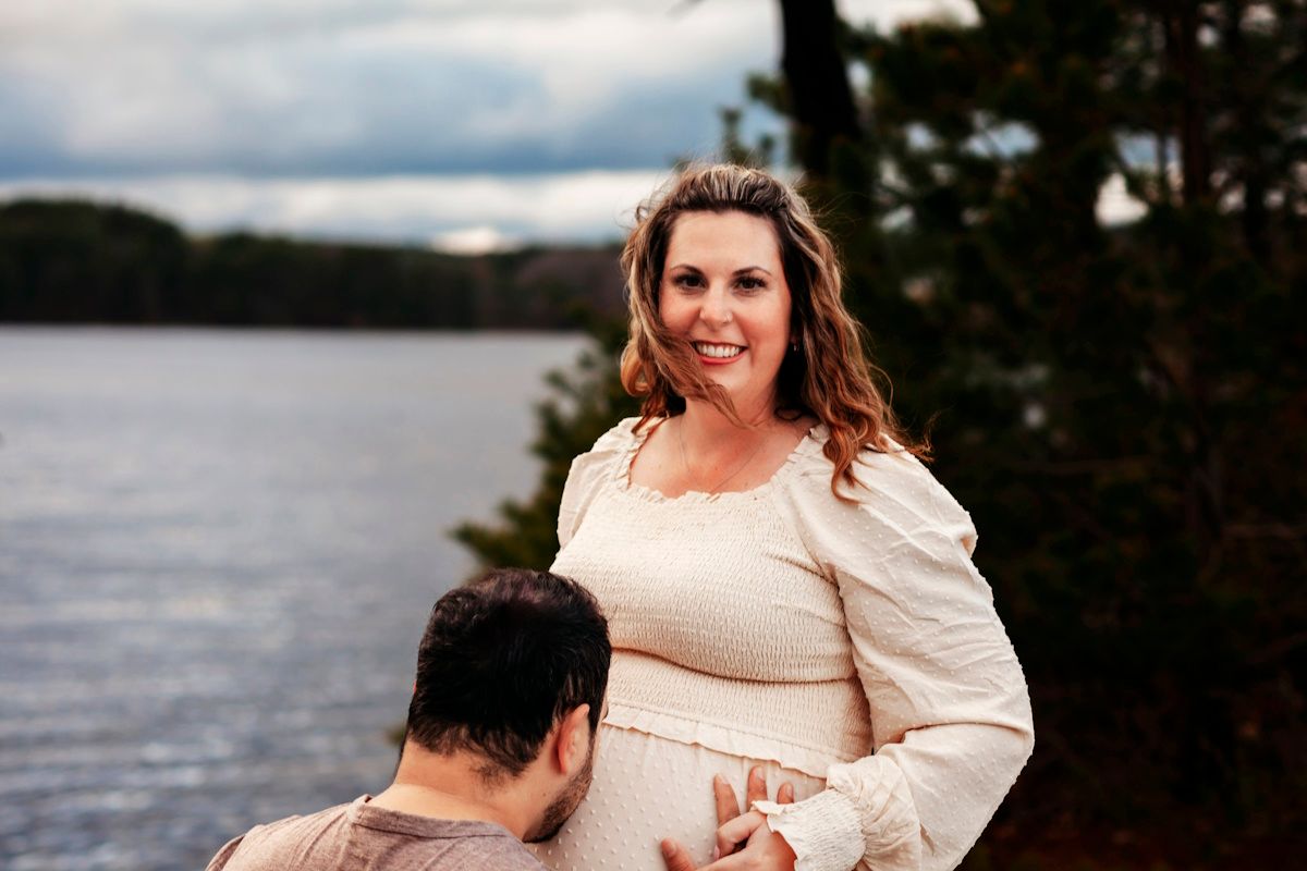 Pregnant woman in cream dress poses with partner by a lake during maternity photo shoot outdoors.