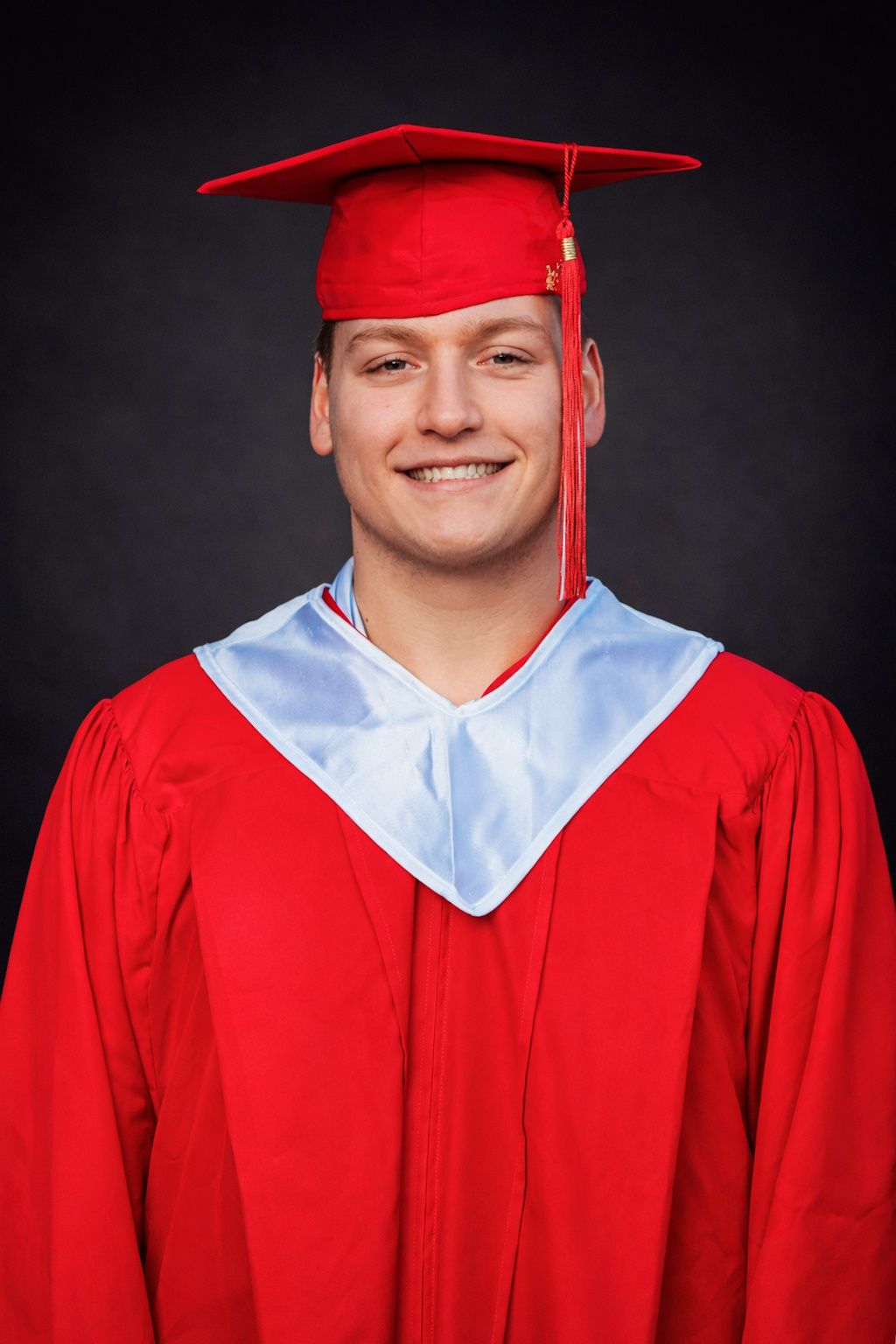 Smiling young man in red graduation cap and gown with light blue collar against dark background.