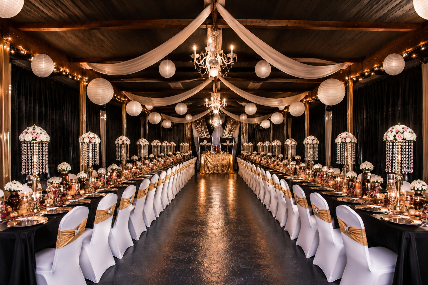 Elegant wedding reception hall with long tables, white chair covers, gold accents, paper lanterns, and draped fabric ceiling decor.