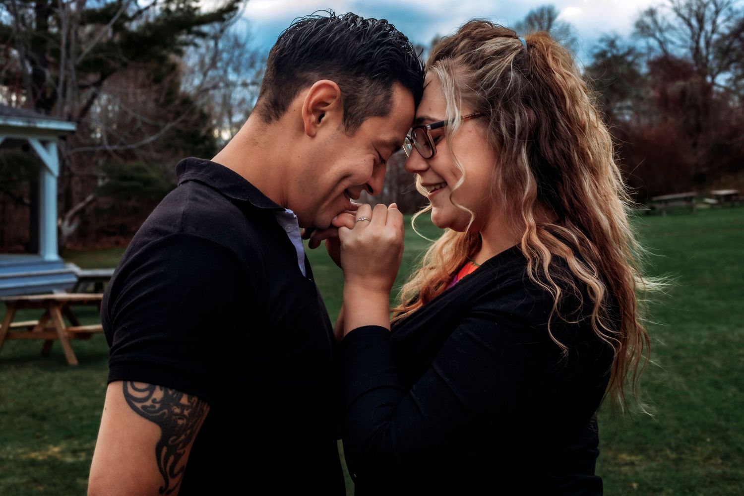 Couple sharing a romantic moment outdoors, touching foreheads and smiling, man with tattoo sleeve, woman in sunglasses.