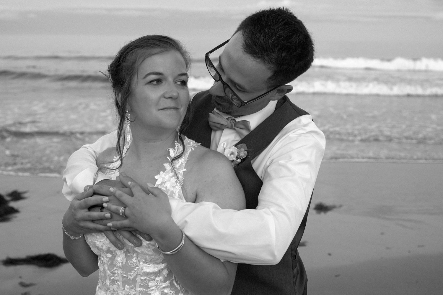 Black and white beach wedding photo of bride and groom embracing by the ocean waves.
