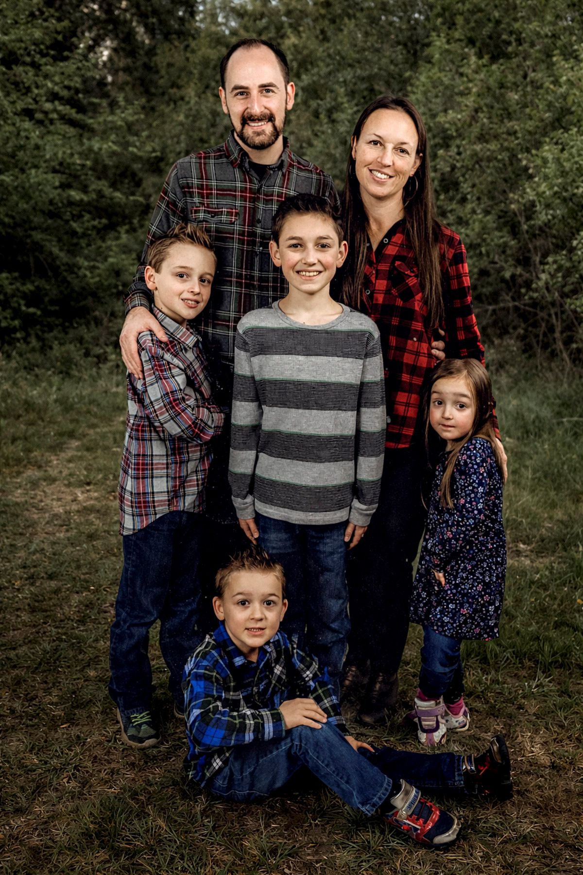 A happy family of six poses outdoors in coordinating plaid and striped outfits, surrounded by lush green trees.