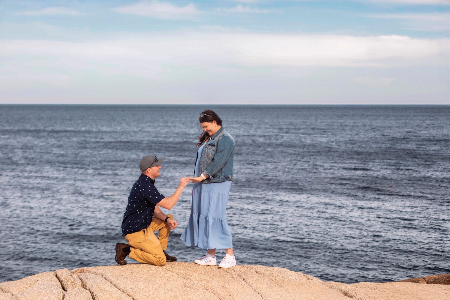 in Halifax Man kneeling on rocky coastal cliff proposing to woman with ocean horizon backdrop on sunny day.