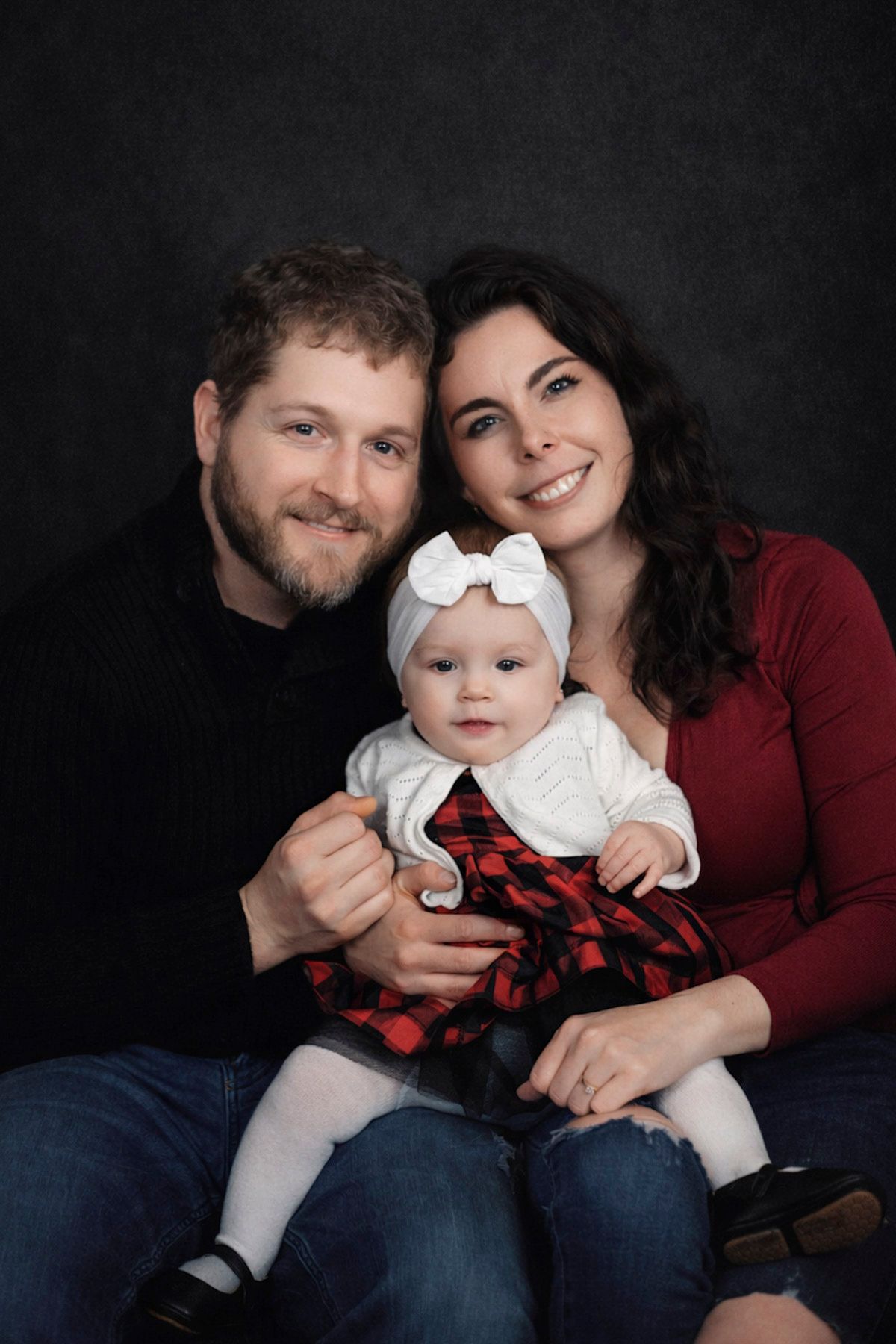 A smiling family portrait featuring a bearded man, dark-haired woman in red, and baby girl in plaid dress with white bow headband.