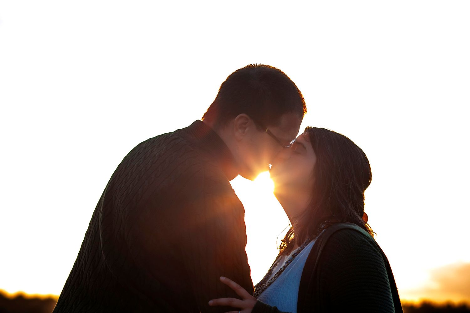 Romantic couple silhouetted against golden sunset light sharing a tender kiss outdoors.