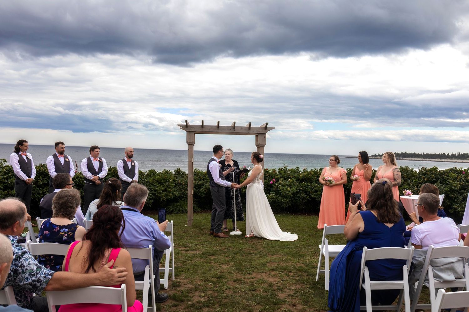 Outdoor waterfront wedding ceremony with couple kissing at wooden arch, groomsmen, bridesmaids in coral dresses, under cloudy sky.