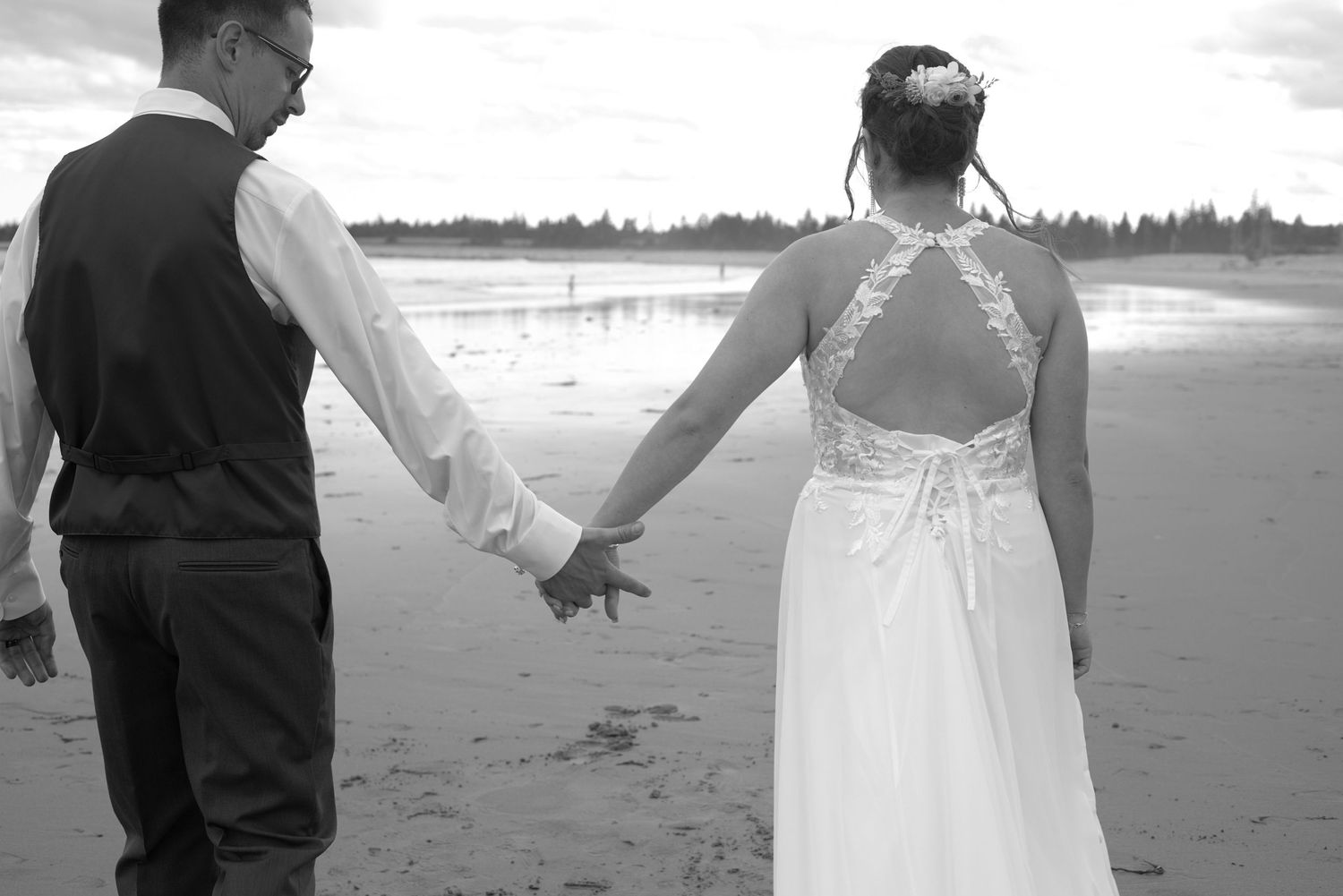 Black and white photo of a bride and groom holding hands on a beach, the bride wearing a lace-back wedding gown.