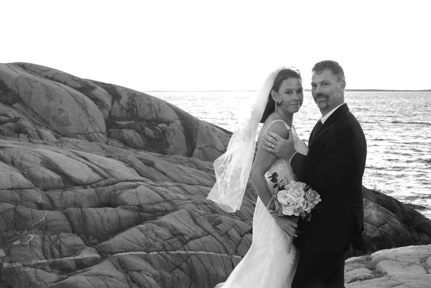 Black and white wedding photo of a bride in a veil and gown holding a bouquet beside a groom in a suit on rocky shoreline by the sea.