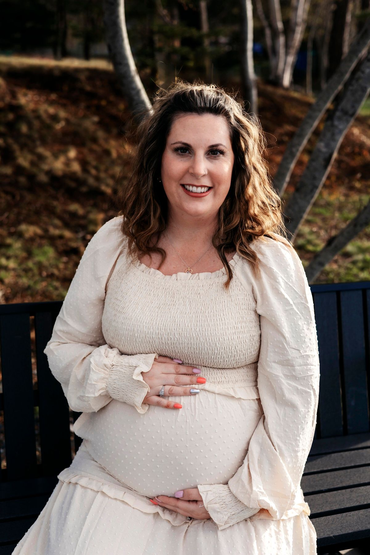 Smiling pregnant woman in cream smocked dress cradling baby bump outdoors on bench with autumn trees background.