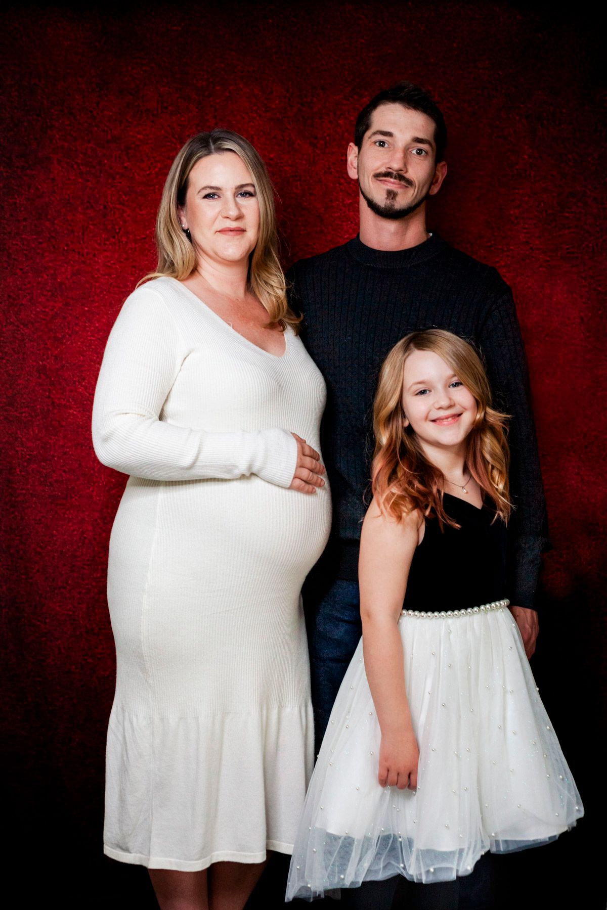 Annapolis Valley Pregnant woman in white dress poses with man in black sweater and young girl in tutu skirt against red backdrop.