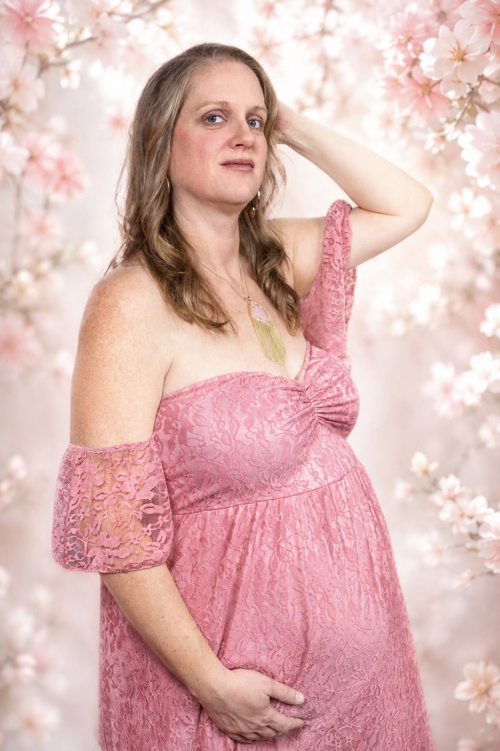Pregnant woman in pink lace off-shoulder dress poses gracefully among cherry blossoms in a soft floral studio setting.
