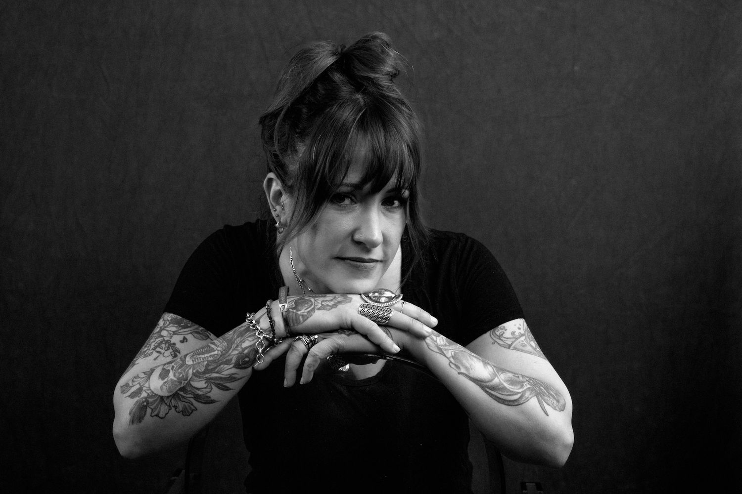 Black and white portrait of a tattooed woman in black top, hands clasped, looking down thoughtfully.