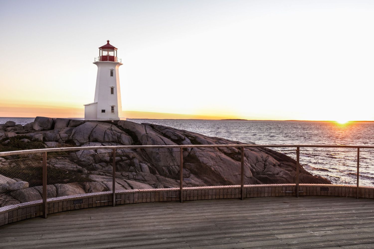 White lighthouse with red top on rugged coastal rocks at sunset, viewed from a wooden observation deck overlooking the calm ocean.