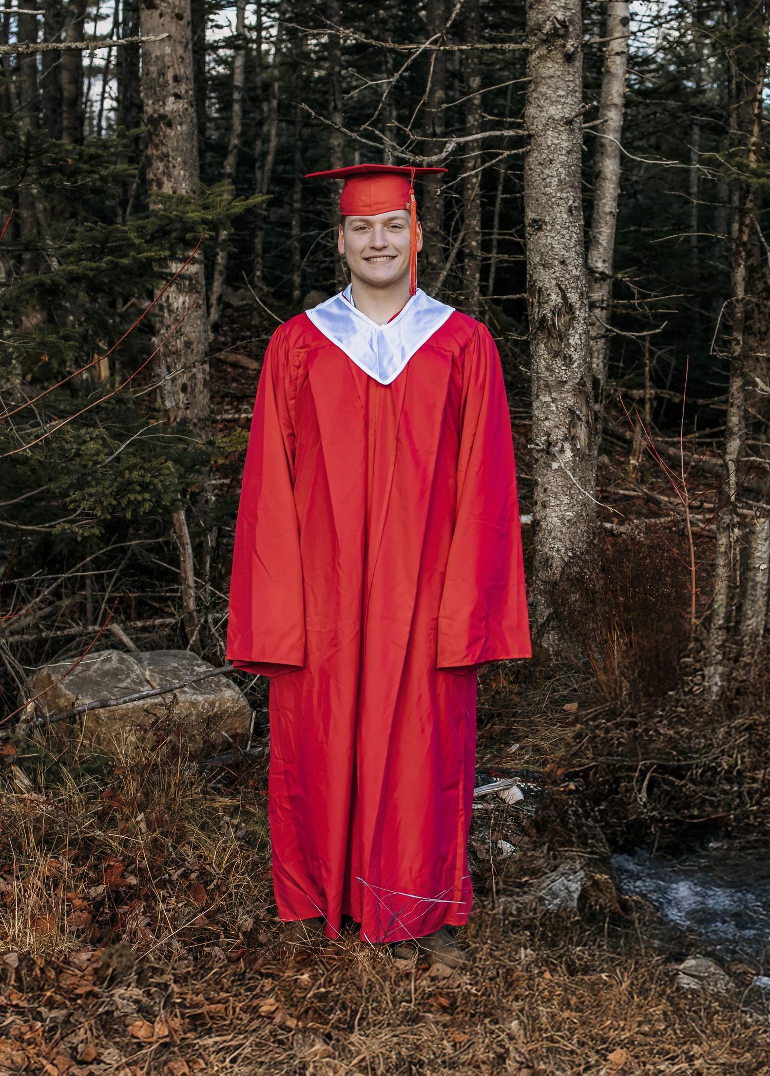 Young man in red graduation cap and gown smiling outdoors in a wooded setting with fallen leaves.