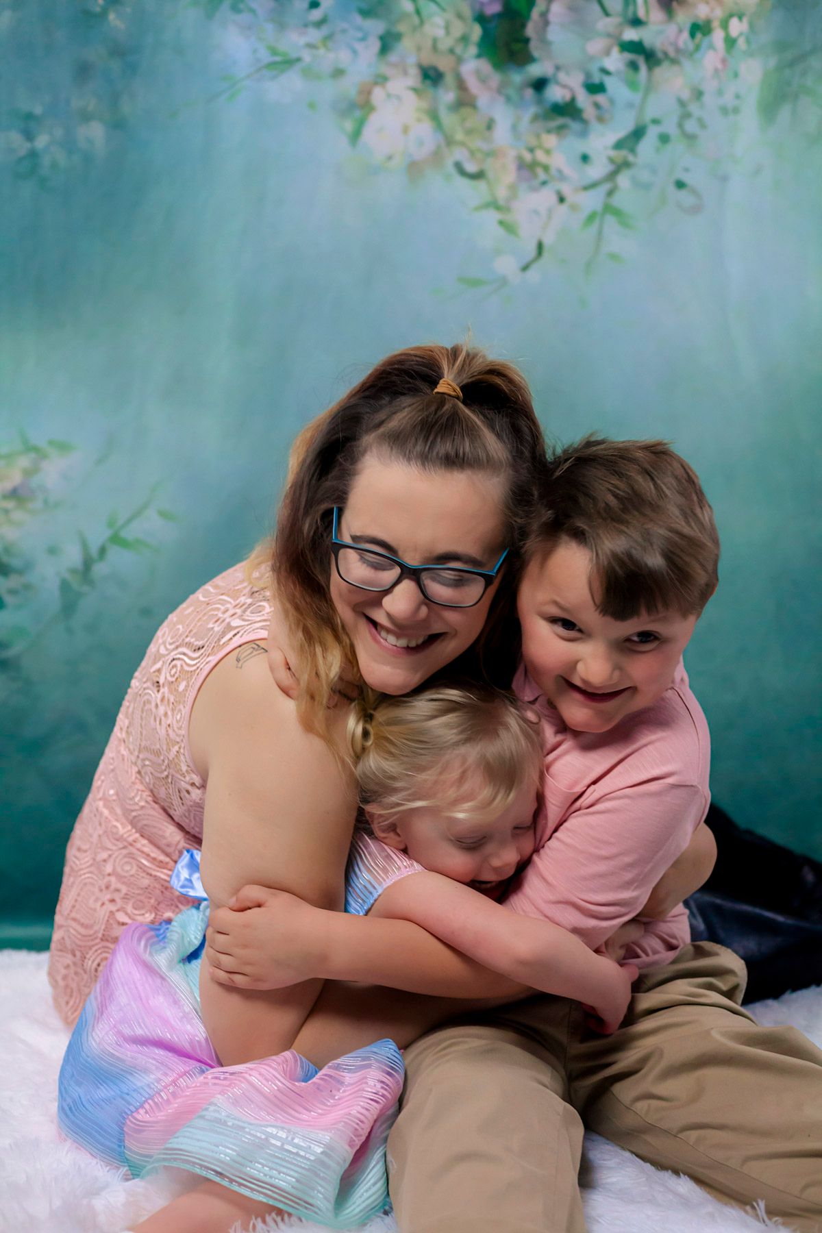 Annapolis family, A smiling woman with glasses hugs two young children against a floral teal studio backdrop.