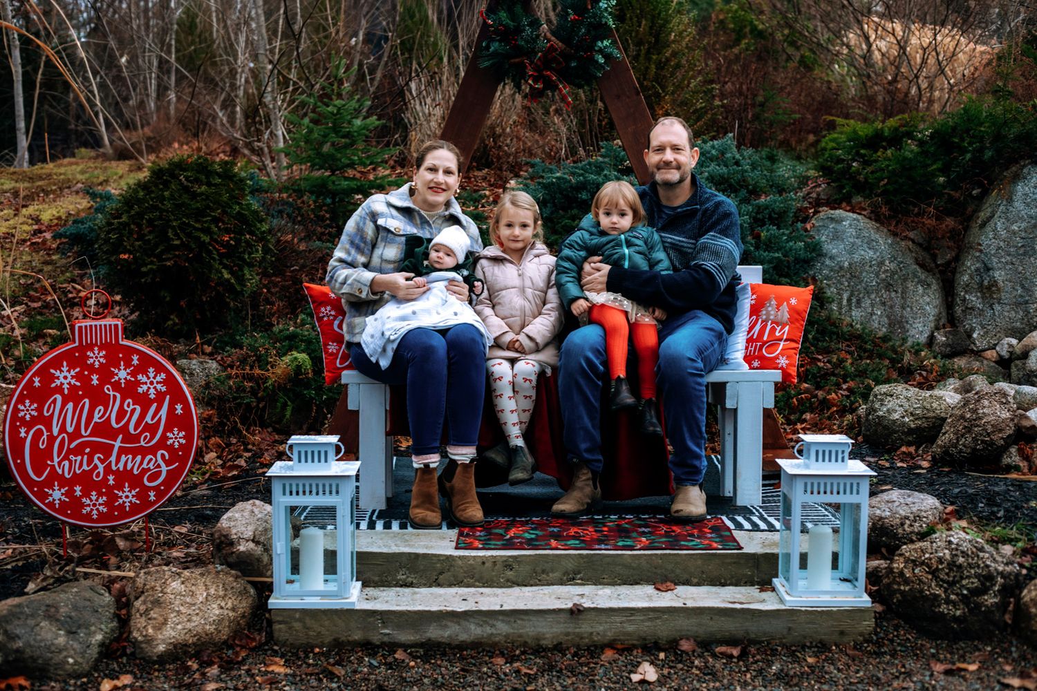 Family of five sits on a white bench outdoors beside rocks and white lanterns, with a red Merry Christmas sign for a holiday photo.