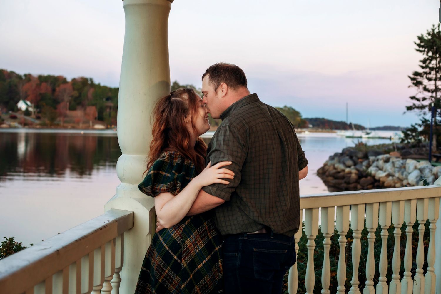 Couple sharing a romantic kiss on a waterfront balcony at dusk with scenic harbor views.