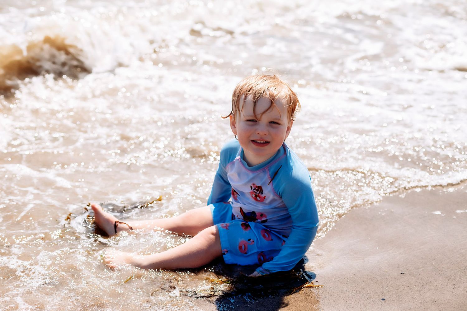 Toddler in blue rash guard sitting in ocean waves on sandy beach, smiling and enjoying the water.