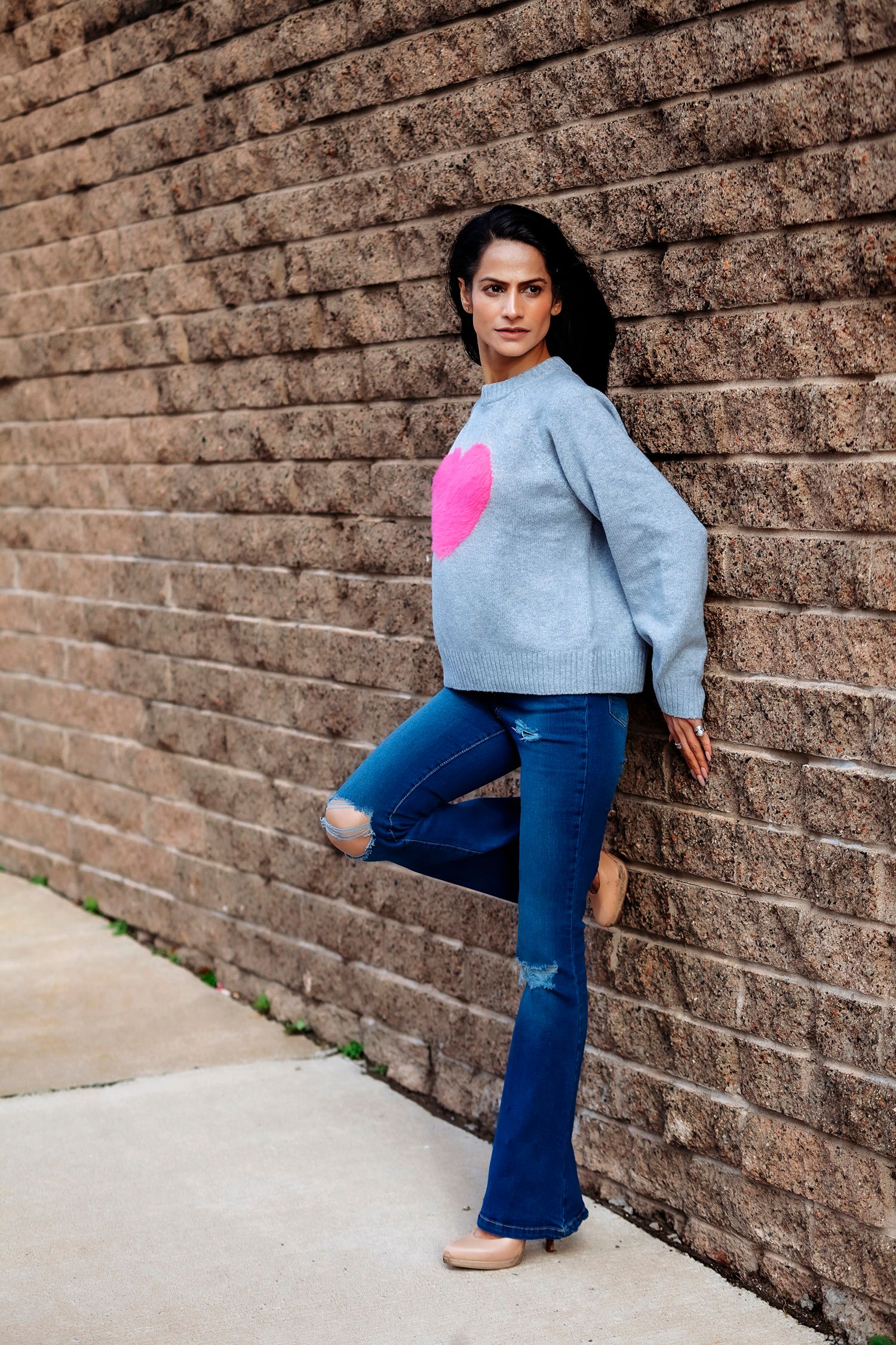 Woman in gray heart sweater and flare jeans posing against brick wall.