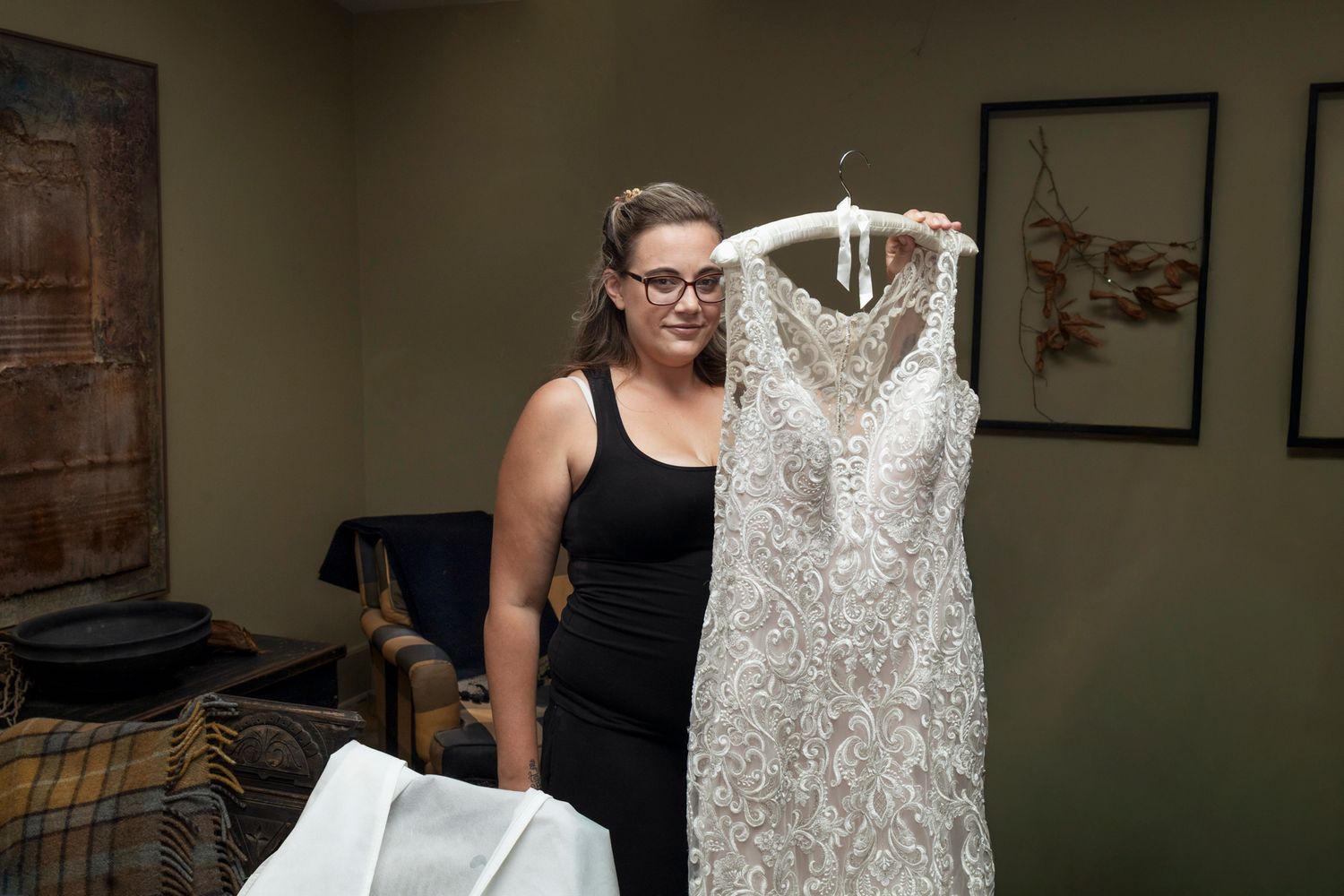 Woman in black dress holding an elegant beaded white wedding gown on a hanger in a softly lit room.