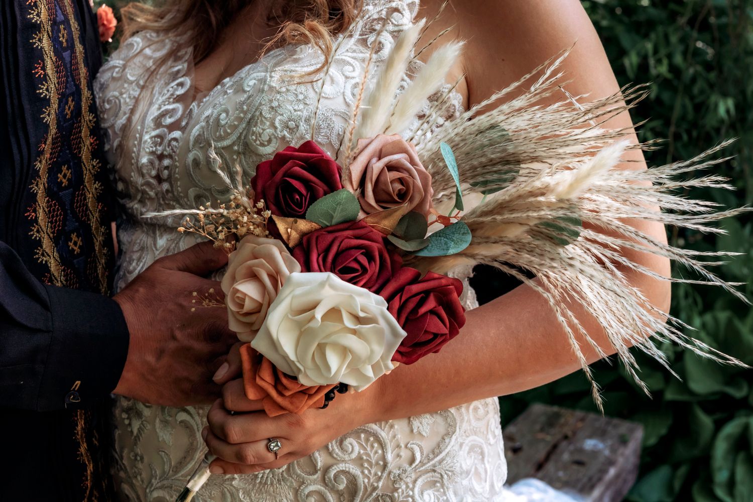 Bride holding a boho wedding bouquet with red roses, blush flowers, and dried pampas grass in a lace gown.