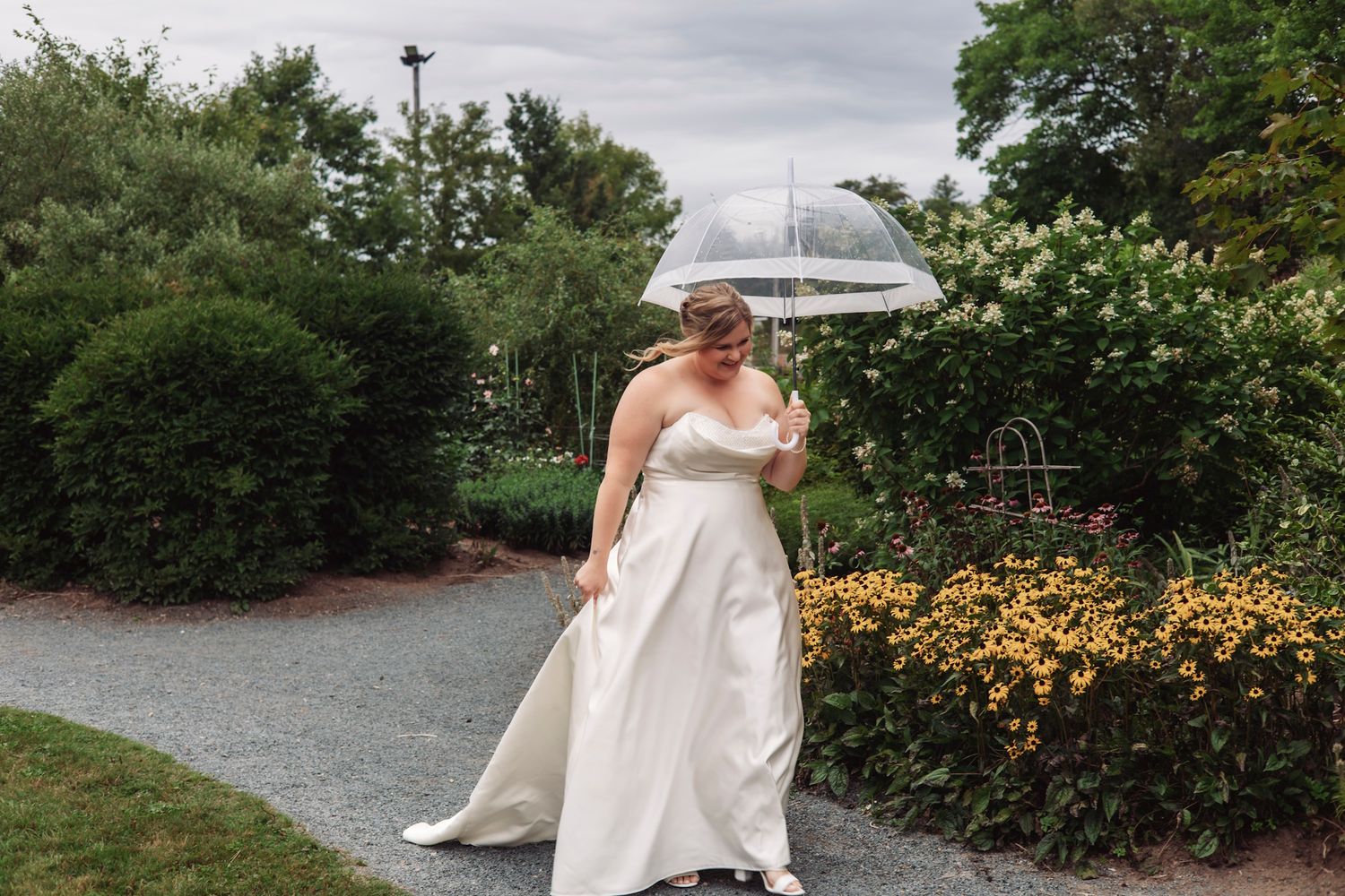 Bride in white gown holding clear umbrella walks through lush garden path with yellow flowers on cloudy wedding day.