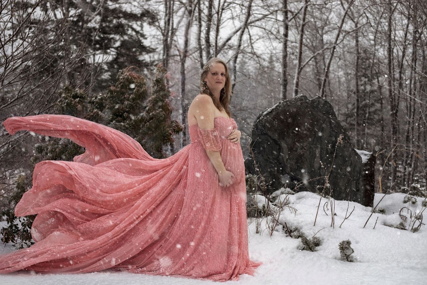 Pregnant woman in flowing pink gown standing in snowy winter forest during snowfall.