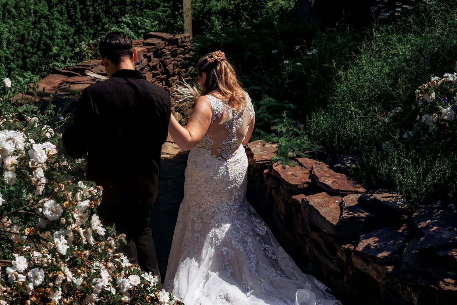 Bride in lace wedding gown and groom in black suit walk hand in hand through a lush garden with stone wall.