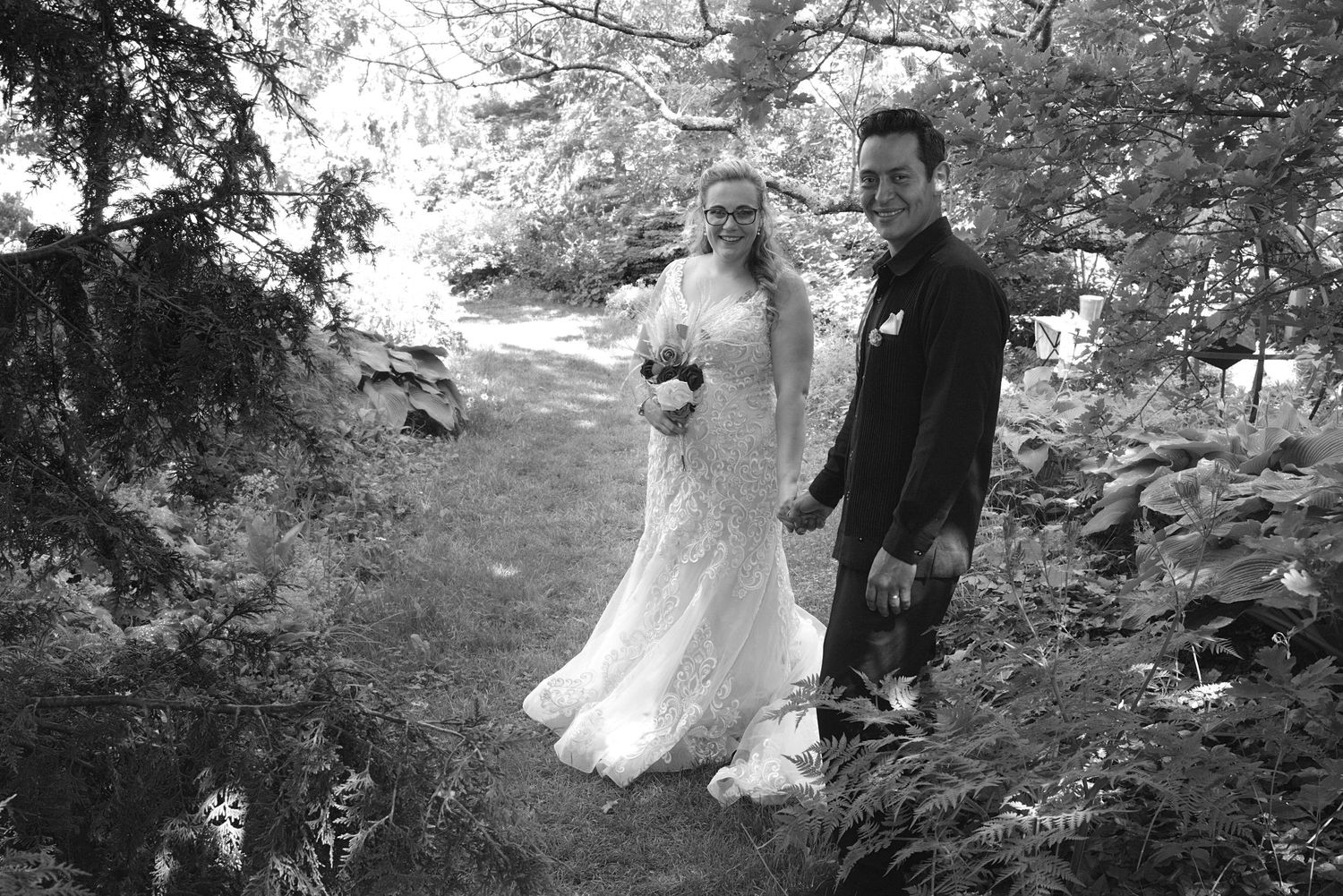 Black and white photo of a bride and groom walking hand in hand through a lush garden on their wedding day.