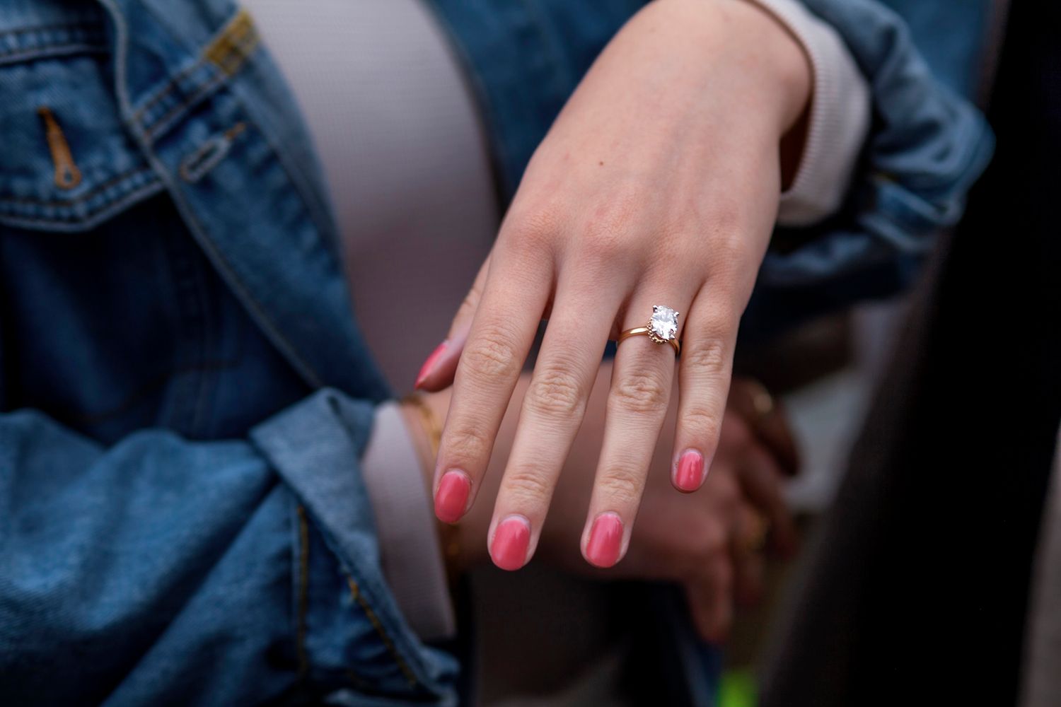 in Halifax a Close-up of a woman's hand with pink nails wearing a diamond engagement ring over a denim jacket.