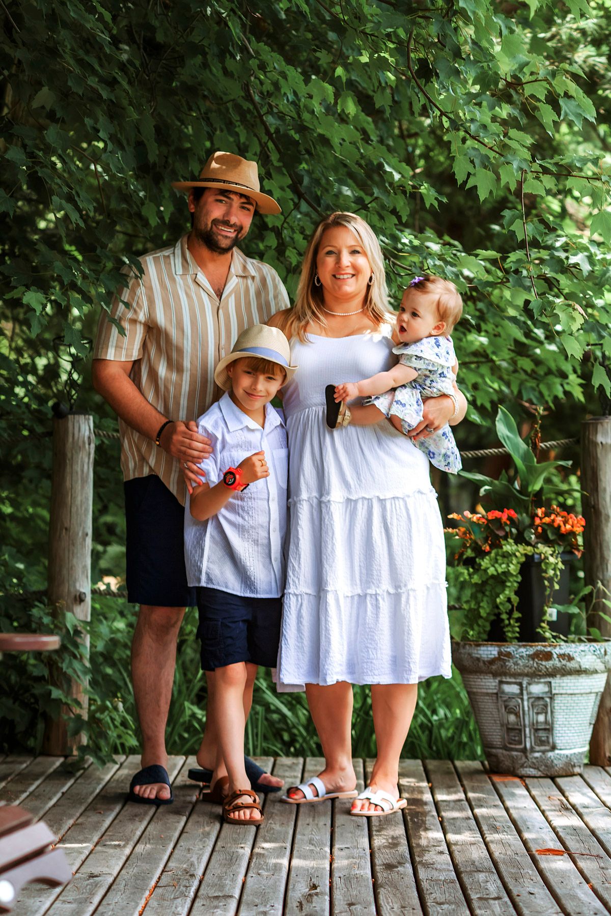 A smiling family of four poses on a wooden deck surrounded by lush green trees, with parents holding two young children.