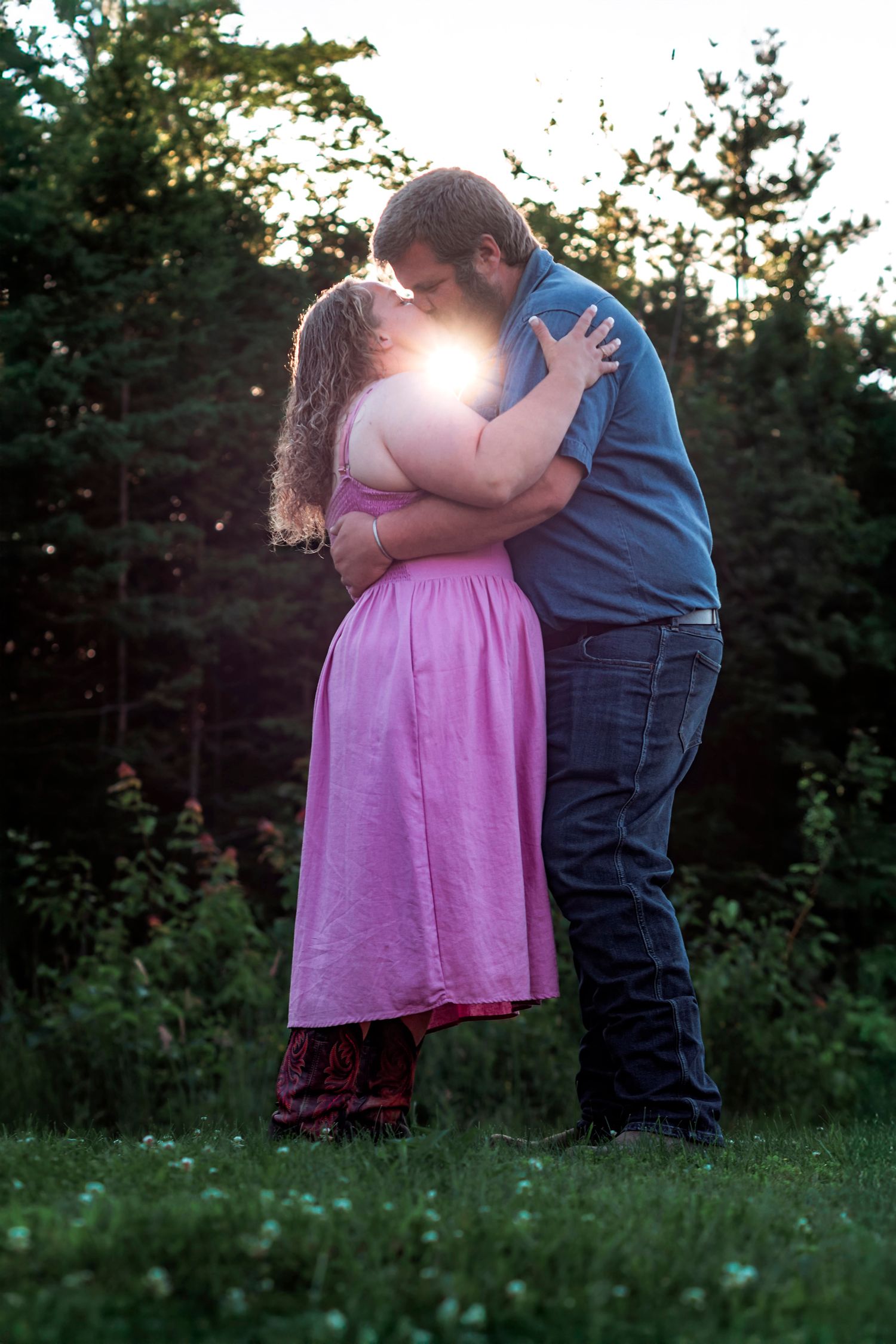 Couple sharing a romantic kiss at sunset in a lush green garden, woman in pink dress embraced by man in blue shirt.
