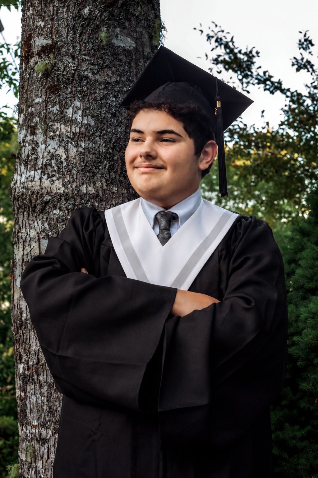 Young graduate in black cap and gown leans against tree with arms crossed, smiling outdoors at the Atlantic Memories Graduation sessions