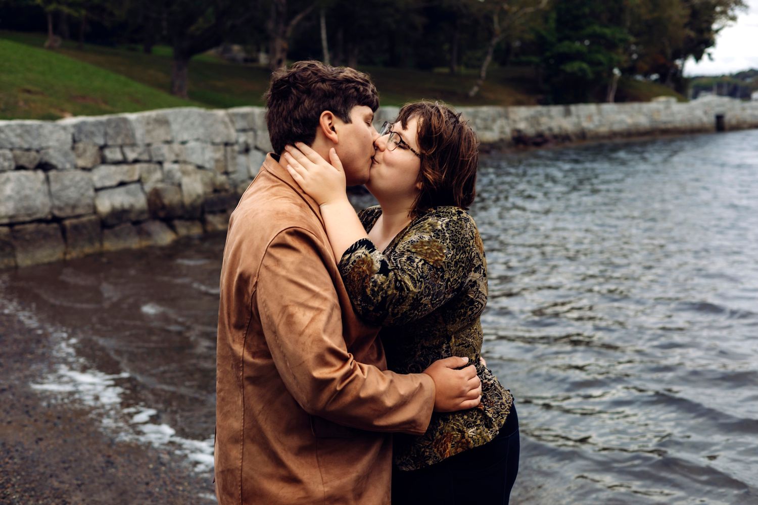 Couple sharing a romantic kiss by a rocky waterfront, woman in floral top embraced by man in brown shirt.