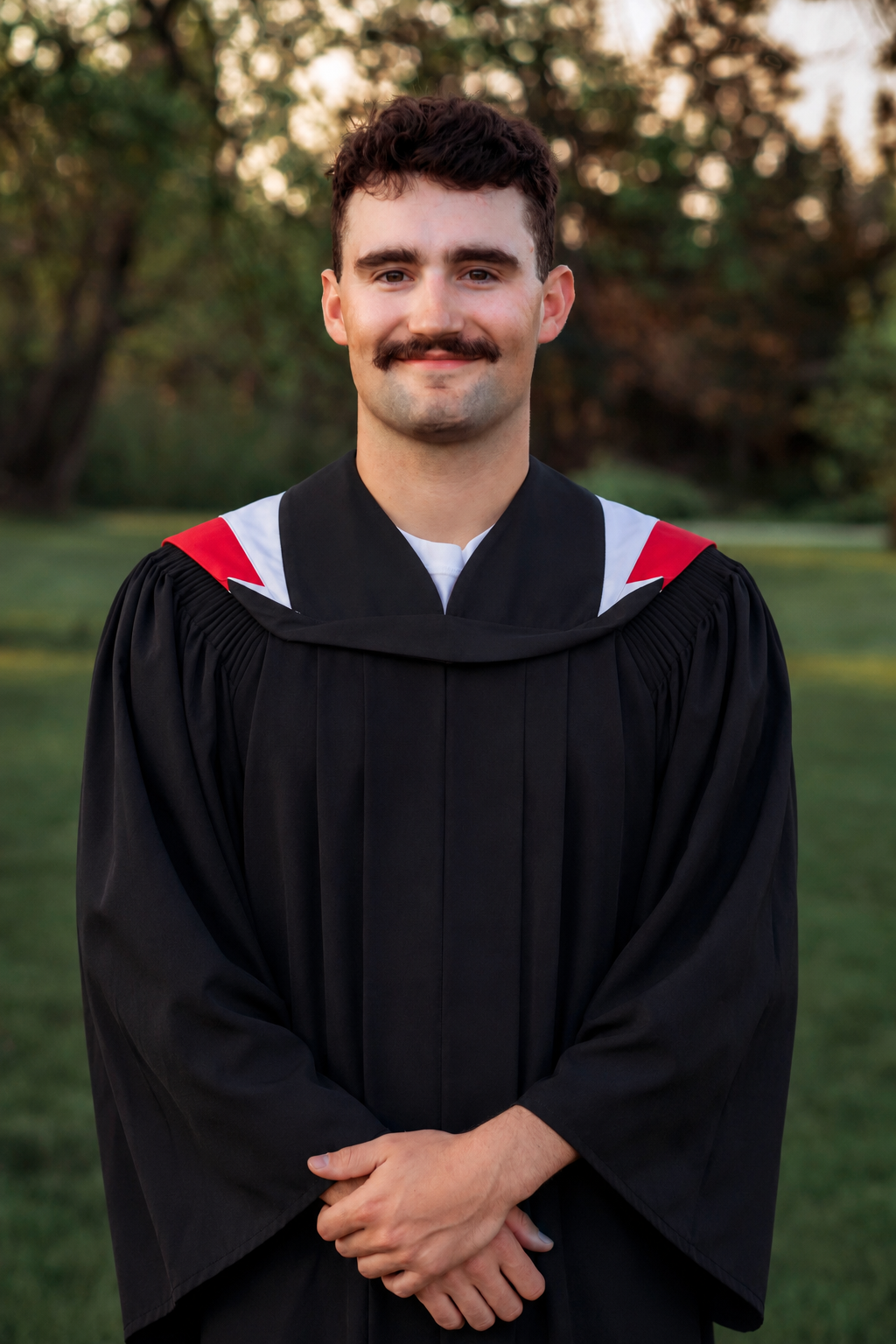 Young man with mustache smiling in black graduation gown with red and white hood outdoors.