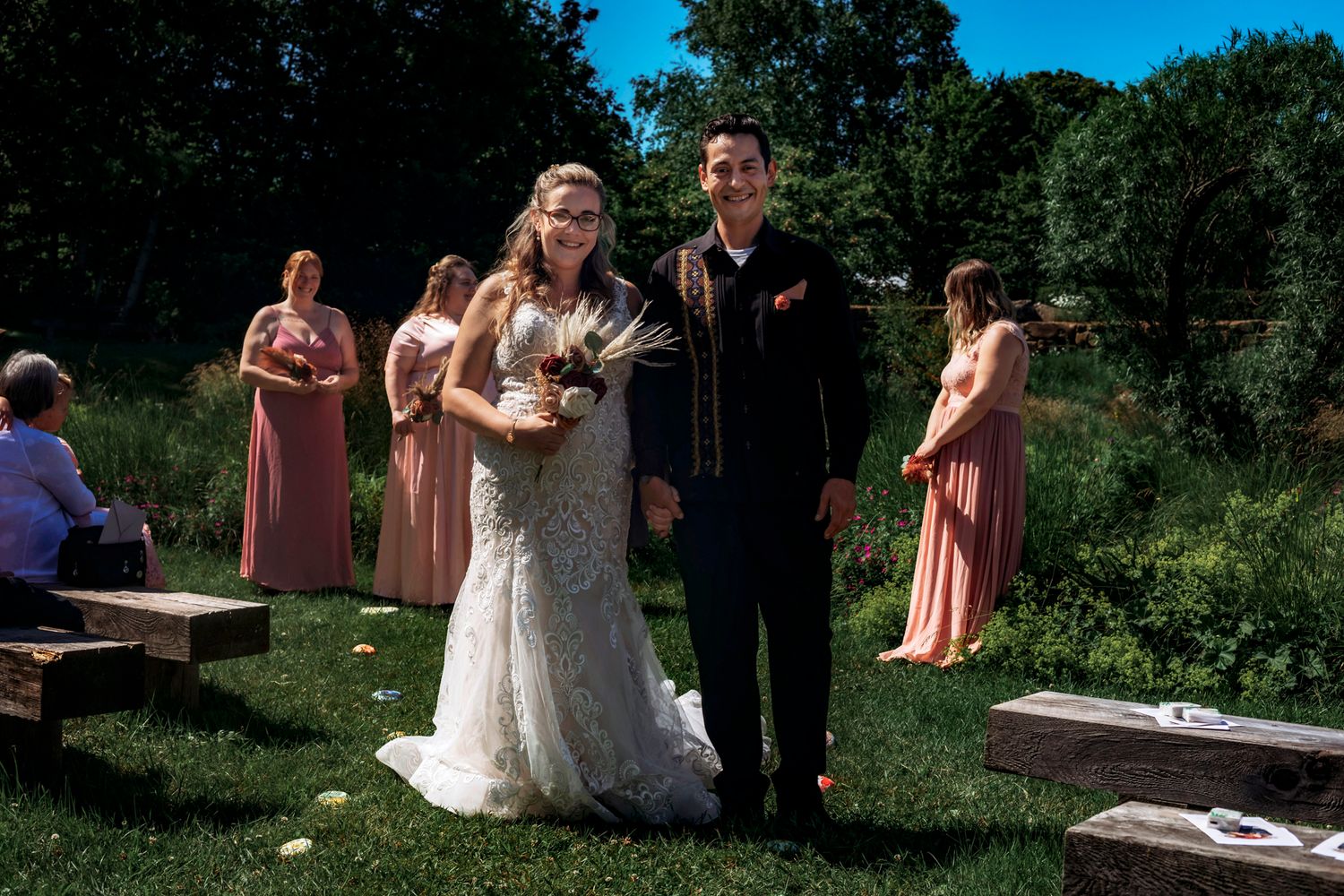 Bride and groom walk down outdoor aisle at garden wedding, bridesmaids in pink dresses standing behind them.