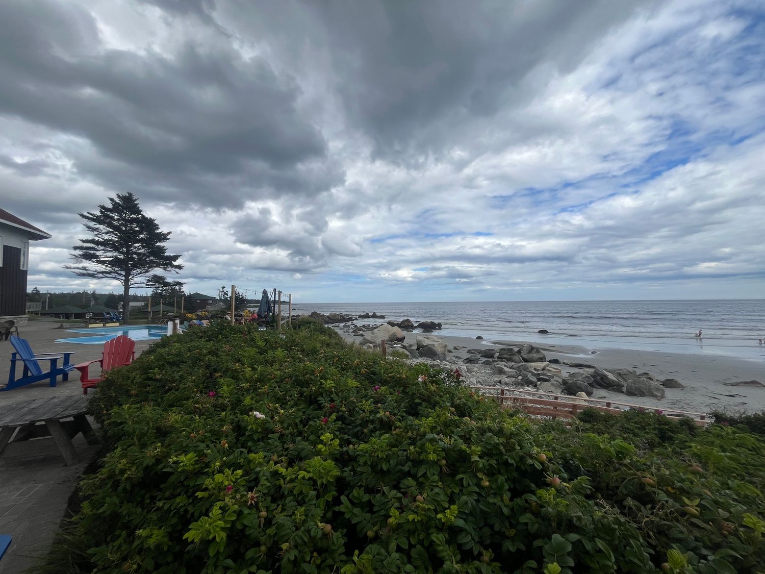 Cloudy coastal scene with rocky beach, lush greenery, Adirondack chairs, and ocean views under dramatic skies.