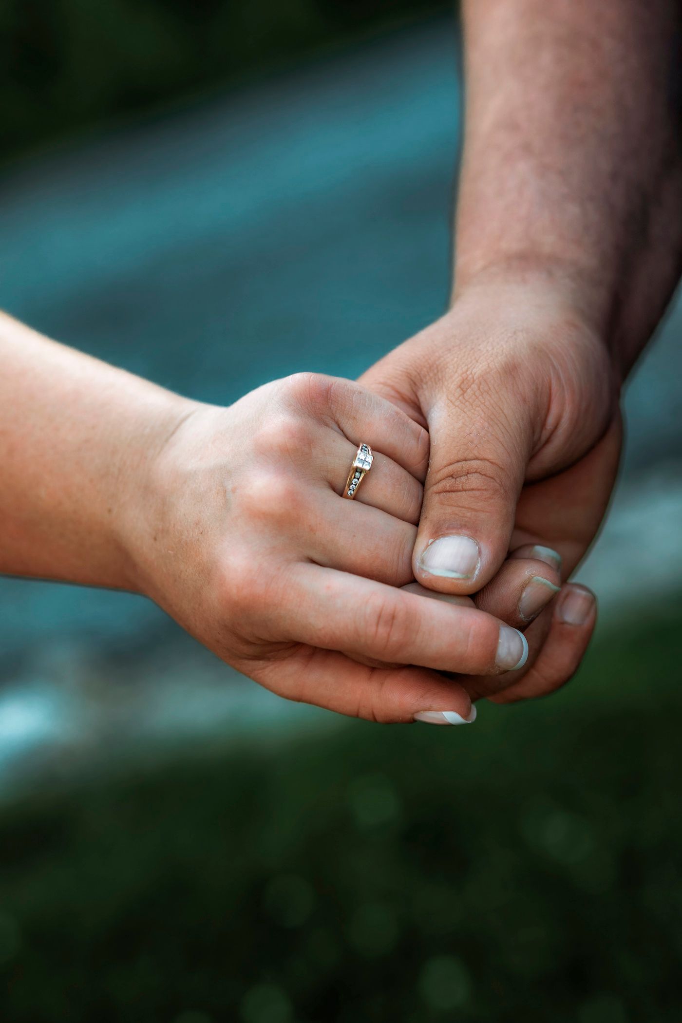 Two hands holding together, one wearing a gold diamond engagement ring, against a blurred green and blue background.