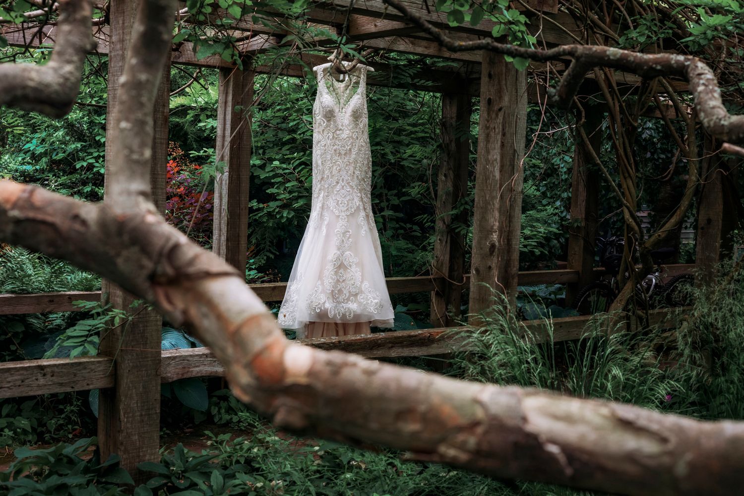 A lace wedding dress hanging from a rustic wooden arbor surrounded by lush green foliage in an outdoor garden setting.