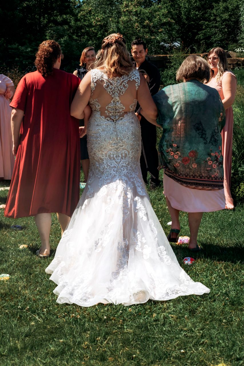 Bride in lace backless wedding gown stands with wedding guests on green lawn during outdoor ceremony.