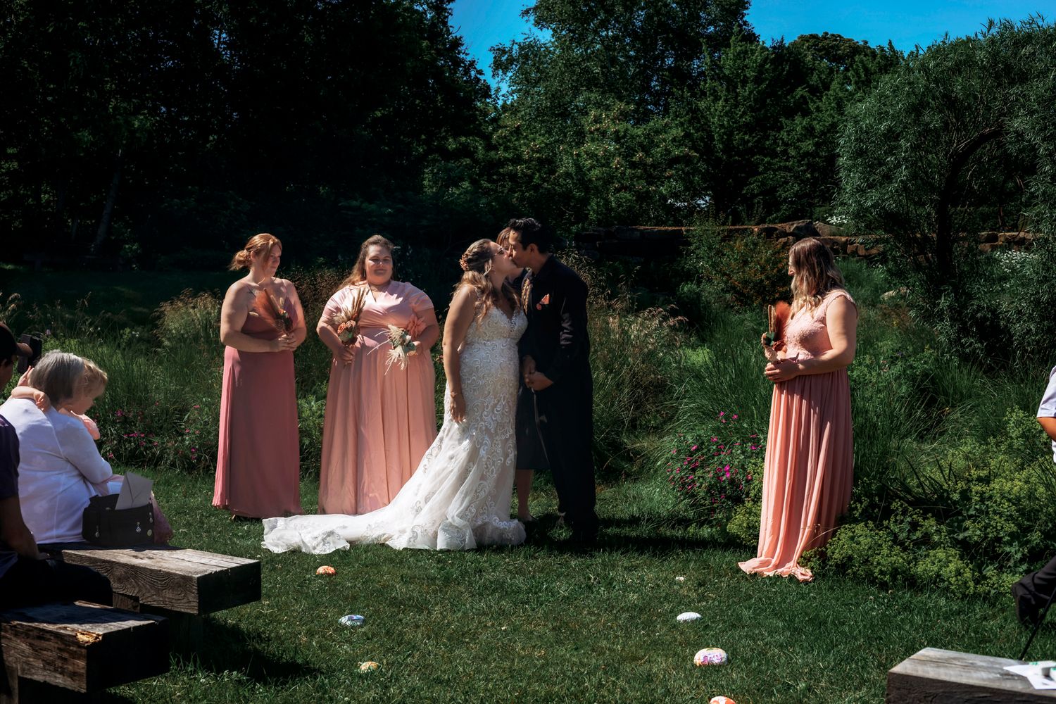 Bride and groom share first kiss at outdoor wedding ceremony with bridesmaids in pink dresses on sunny day.