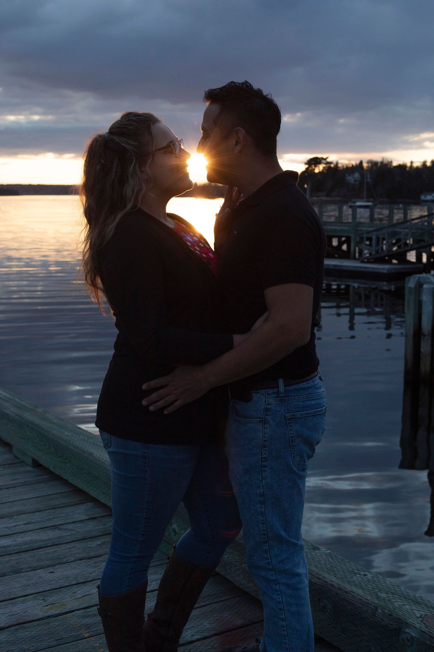 Romantic couple embracing on a waterfront dock at sunset, silhouetted against a dramatic cloudy sky.