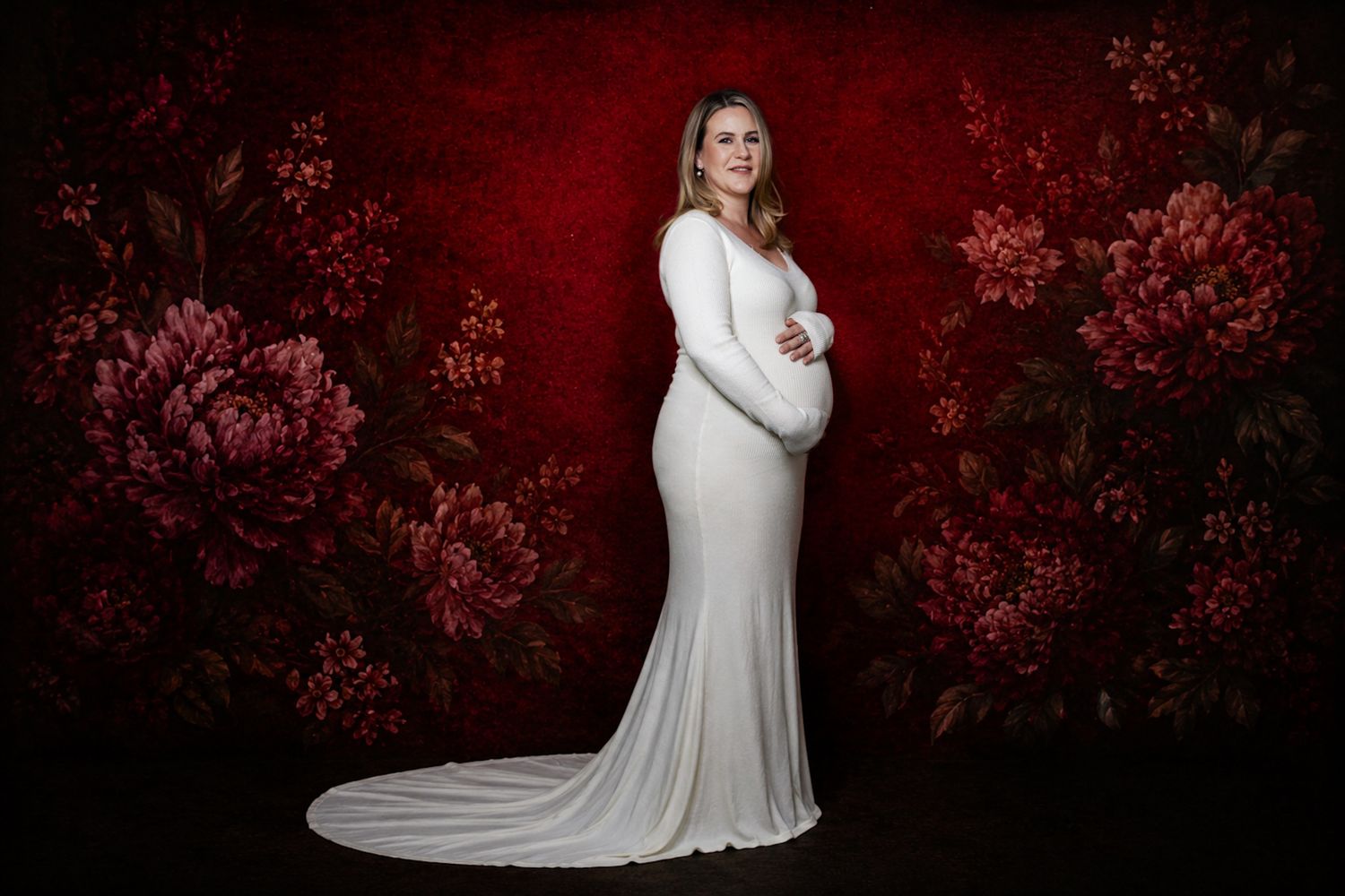 Pregnant woman in white fitted gown posing against red floral backdrop in elegant maternity portrait photography.