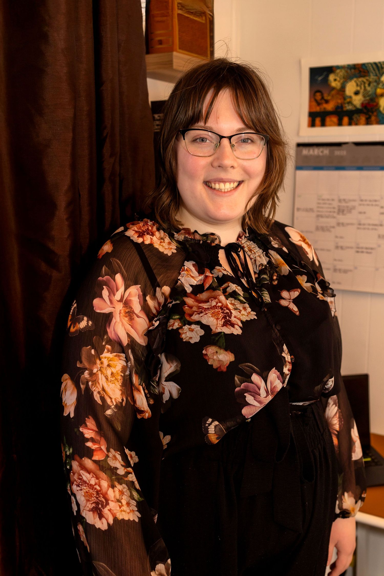 Young woman with glasses smiling in a black floral dress, standing indoors near a calendar on the wall.