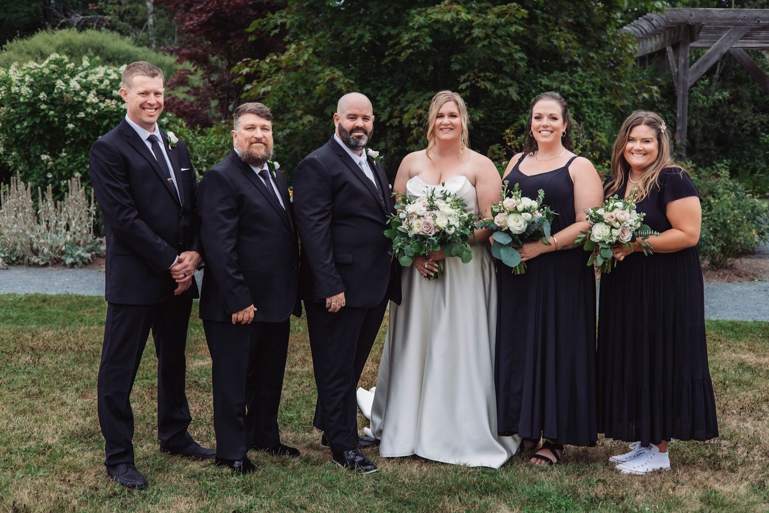Wedding party posing outdoors, bride in white gown with bouquet, bridesmaids in black dresses, groomsmen in dark suits.