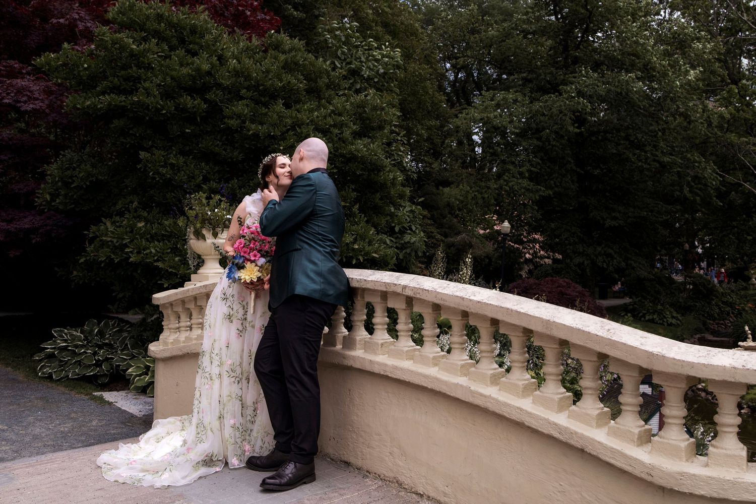Bride and groom share a romantic embrace on an elegant stone balcony surrounded by lush green trees.